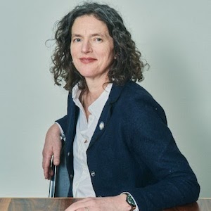 image of smiling white woman with dark curly hair sitting at a table wearing dark blue blazer and white button up shirt